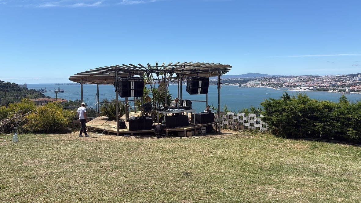 A timber-canopied stage on an open grass hilltop, speakers rigged overhead, with the Tagus estuary and Lisbon on the far bank
