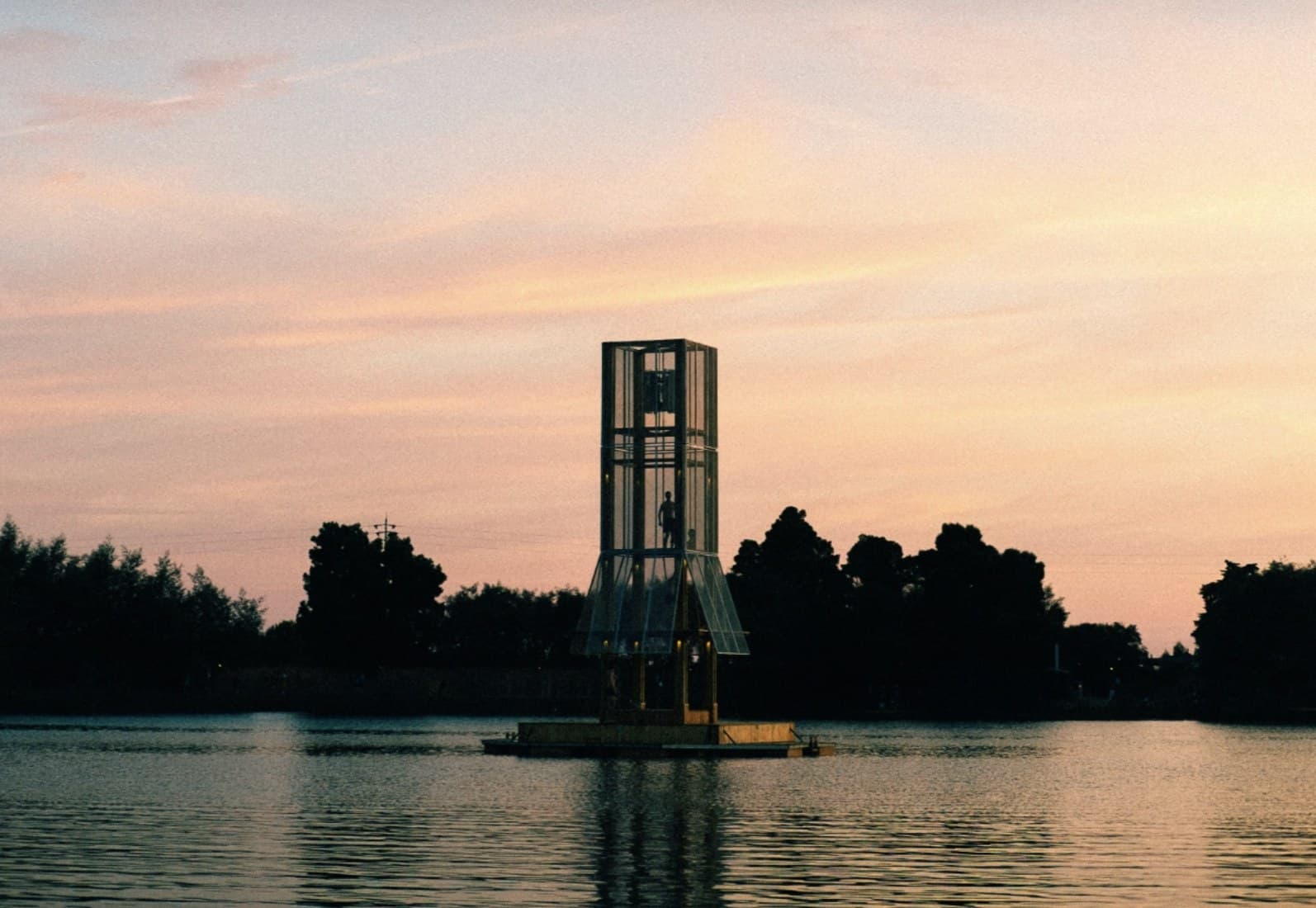 A glazed timber tower on a floating pontoon at dusk, a single figure standing inside it, pine woods dark along the far shore