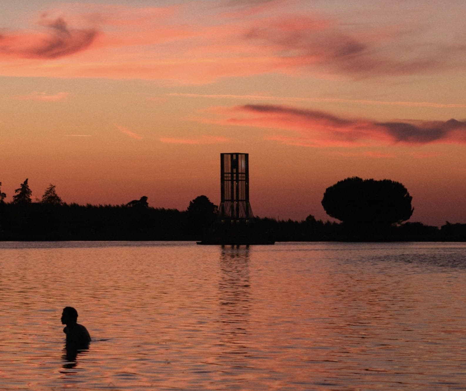 The tower silhouetted against a burning sunset, a swimmer's head and shoulders in the foreground water