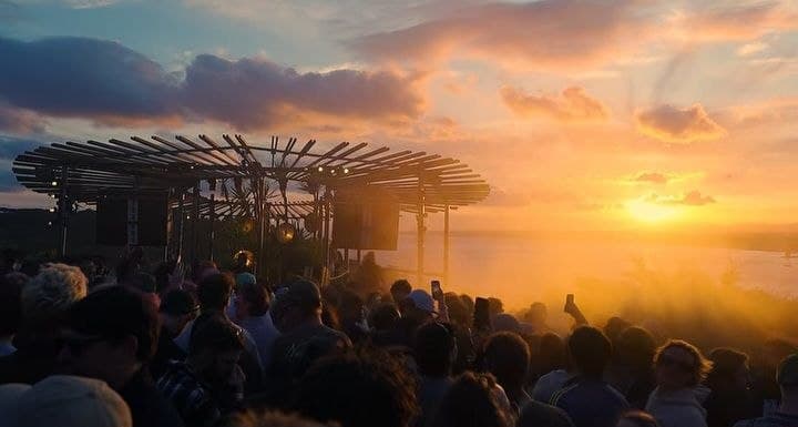 The same stage at sunset, its canopy silhouetted above a dense crowd, low sun burning across the estuary behind it